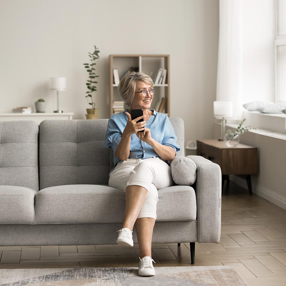 In fashionable cozy living room mature woman sit on sofa with smartphone, looks away, exchanging messages with family